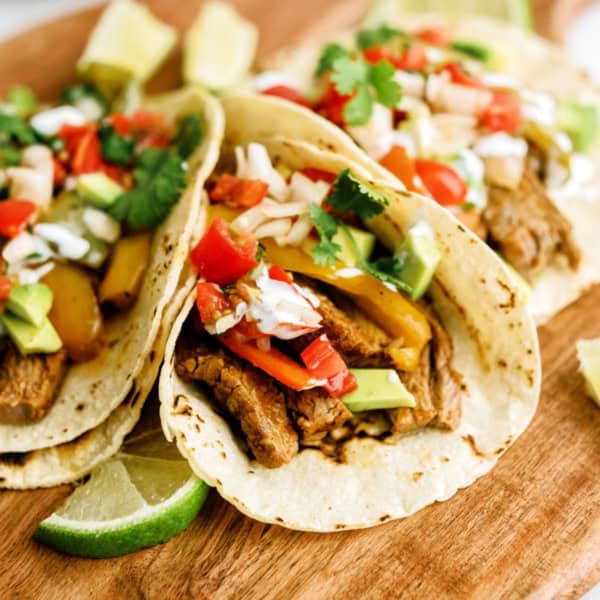 Three loaded tacos with seasoned meat, diced tomatoes, avocado, and cilantro on a wooden board, with lime wedges on the side.