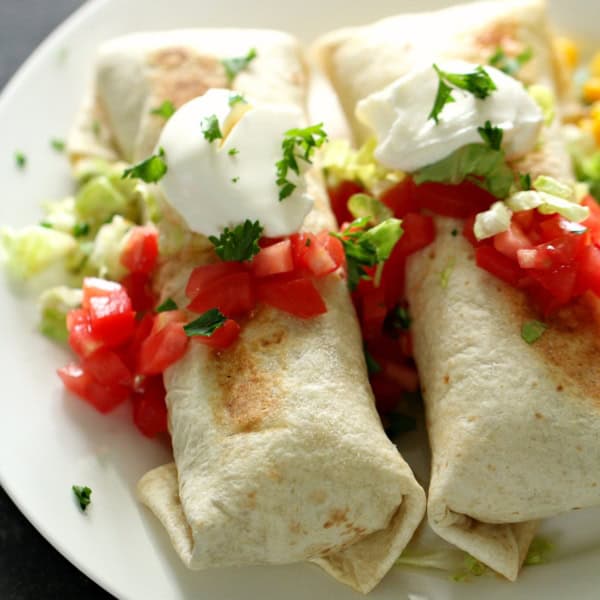Two burritos topped with sour cream and chopped cilantro, served with diced tomatoes and shredded lettuce on a white plate.