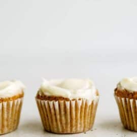 Three frosted muffins in white liners are evenly spaced in a row against a plain white background.