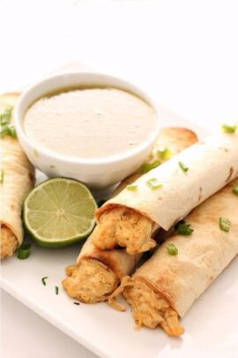 Three chicken taquitos on a plate, served with a lime wedge and a bowl of creamy dipping sauce.