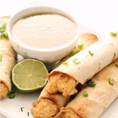 Three chicken taquitos on a plate, served with a lime wedge and a bowl of creamy dipping sauce.