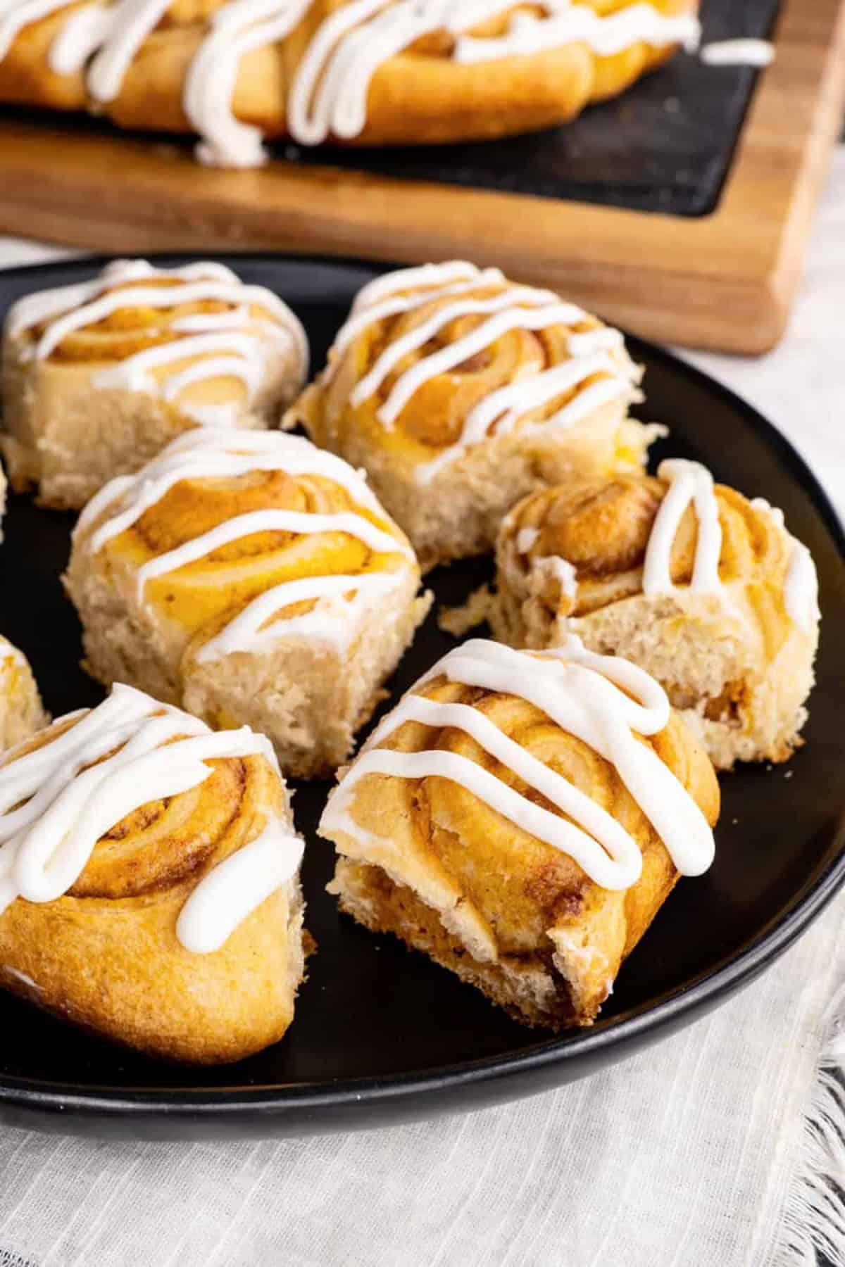 A black plate of cinnamon rolls topped with white icing. More cinnamon rolls on a wooden tray are visible in the background.