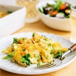 A serving of Chicken and Broccoli Casserole on a white plate with a fork. A salad and remaining casserole in the background.