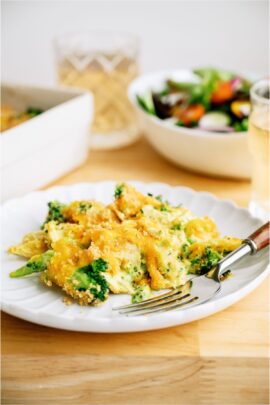 A serving of Chicken and Broccoli Casserole on a white plate with a fork. A salad and remaining casserole in the background.