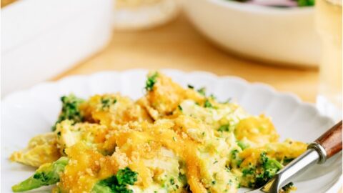 A serving of Chicken and Broccoli Casserole on a white plate with a fork. A salad and remaining casserole in the background.