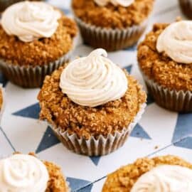 A group of pumpkin cupcakes with crumb topping and swirled frosting arranged on a patterned surface.