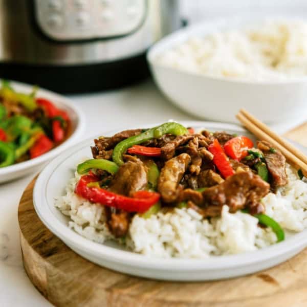 A plate of beef stir-fry with bell peppers on white rice, garnished with chopsticks, with a bowl of rice and vegetables in the background.