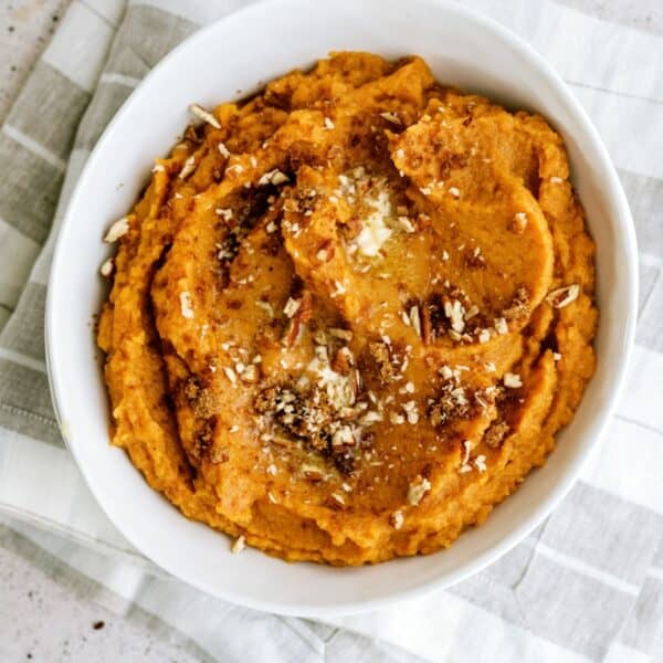 A white bowl filled with mashed sweet potatoes, topped with brown sugar, cinnamon, and chopped nuts, placed on a gray and white striped cloth.