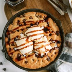 Pizookie in a skillet topped with 3 scoops of ice cream and drizzled with chocolate sauce. Off to the side is a jar or chocolate sauce with a spoon in it and several spoons next to it.