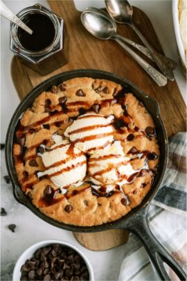 Pizookie in a skillet topped with 3 scoops of ice cream and drizzled with chocolate sauce. Off to the side is a jar or chocolate sauce with a spoon in it and several spoons next to it.