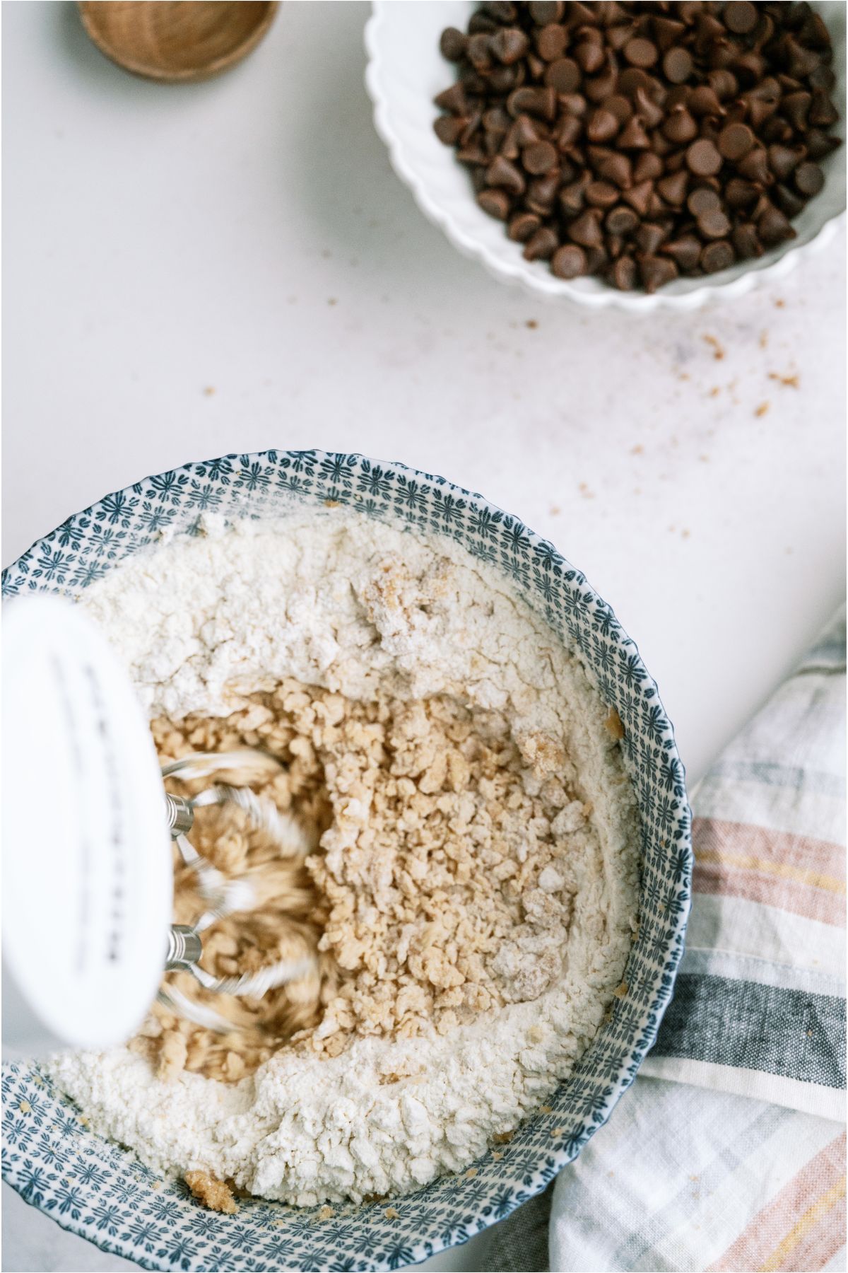 Using a hand mixer to mix dry ingredients into wet ingredients in a mixing bowl. With a bowl of chocolate chips off to the side.