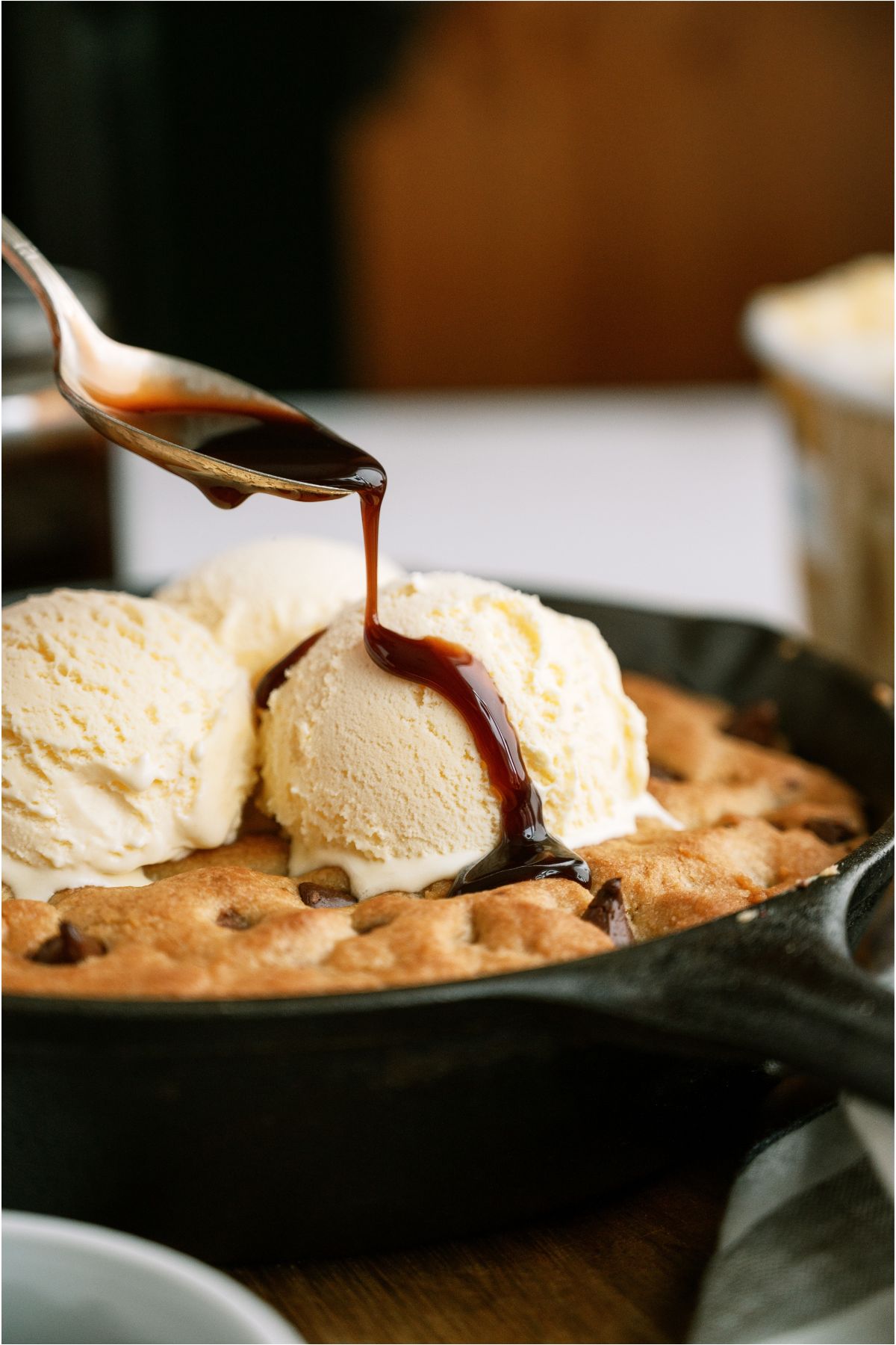 A Pizookie in a skillet with 3 scoops of ice cream on top and a spoon drizzling chocolate sauce on top.