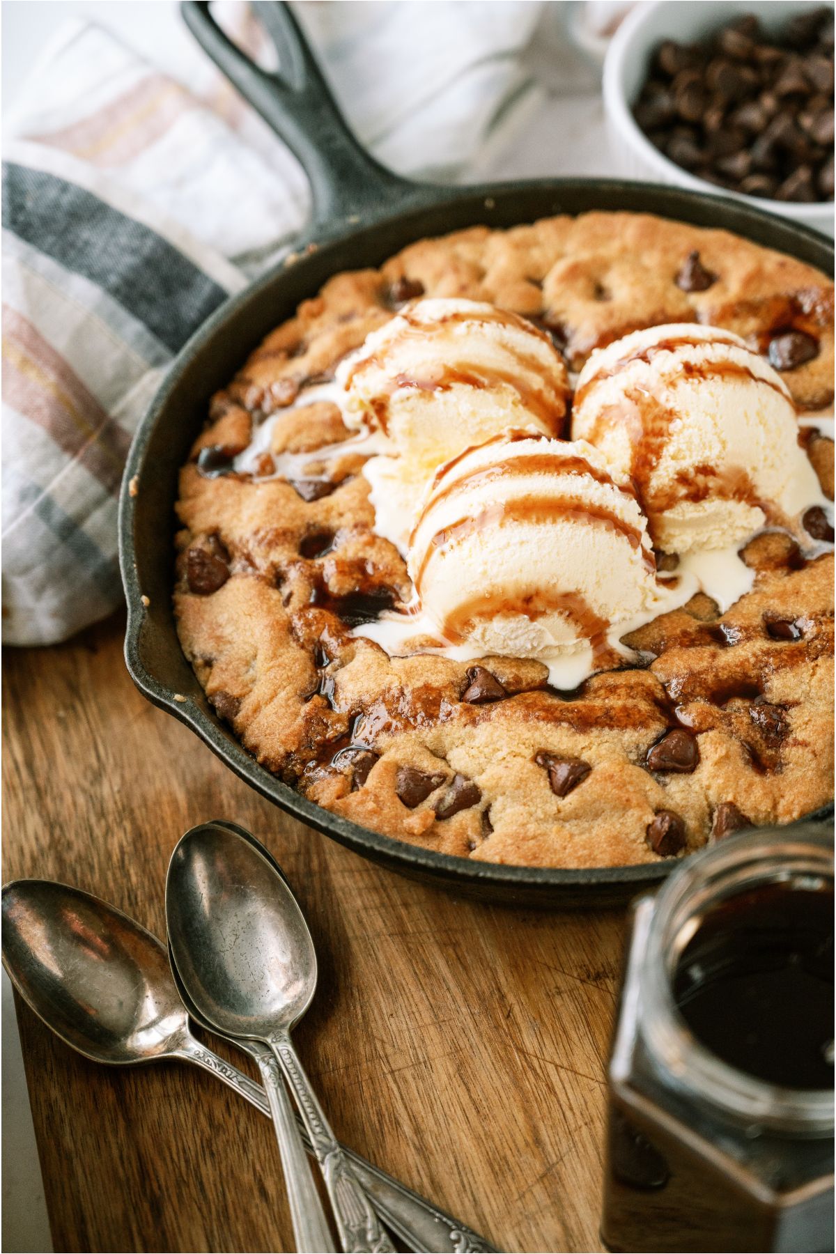 A Pizookie in a skillet with spoons in from and a small jar of chocolate sauce.