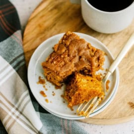 A plate with a slice of pumpkin cake next to a cup of coffee on a wooden board, accompanied by a plaid cloth.