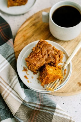 A plate with a slice of pumpkin cake next to a cup of coffee on a wooden board, accompanied by a plaid cloth.