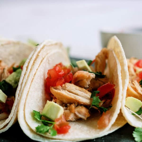 Two tacos filled with shredded meat, diced tomatoes, avocado, and cilantro are arranged on a plate with a bowl visible in the background.
