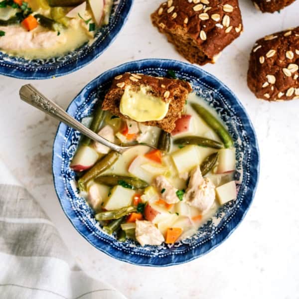 A bowl of vegetable soup with a piece of buttered bread on top. Another piece of bread and a second bowl of soup are in the background.