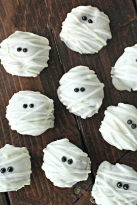 Cookies decorated to resemble mummies with white icing and black eyes, arranged on a wooden surface.