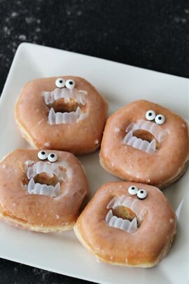 Four glazed donuts are decorated with plastic fangs and candy eyeballs, resembling faces with open mouths and wide eyes, placed on a white square plate.