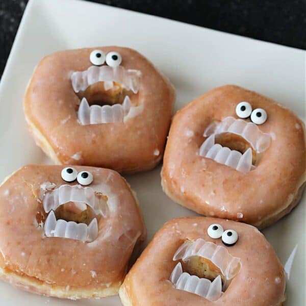 Four glazed donuts are decorated with plastic fangs and candy eyeballs, resembling faces with open mouths and wide eyes, placed on a white square plate.