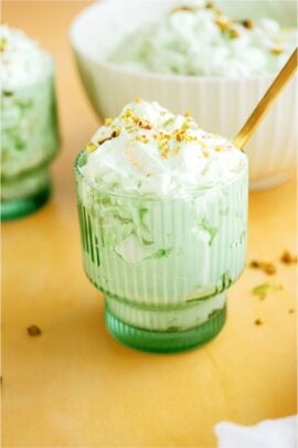 A green glass filled with a serving of Pistachio salad topped with sliced pistachios and a gold spoon. More 5 Minute Pistachio Salad in the background.