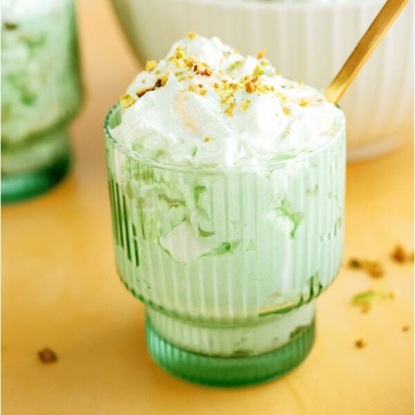 A green glass filled with a serving of Pistachio salad topped with sliced pistachios and a gold spoon. More 5 Minute Pistachio Salad in the background.