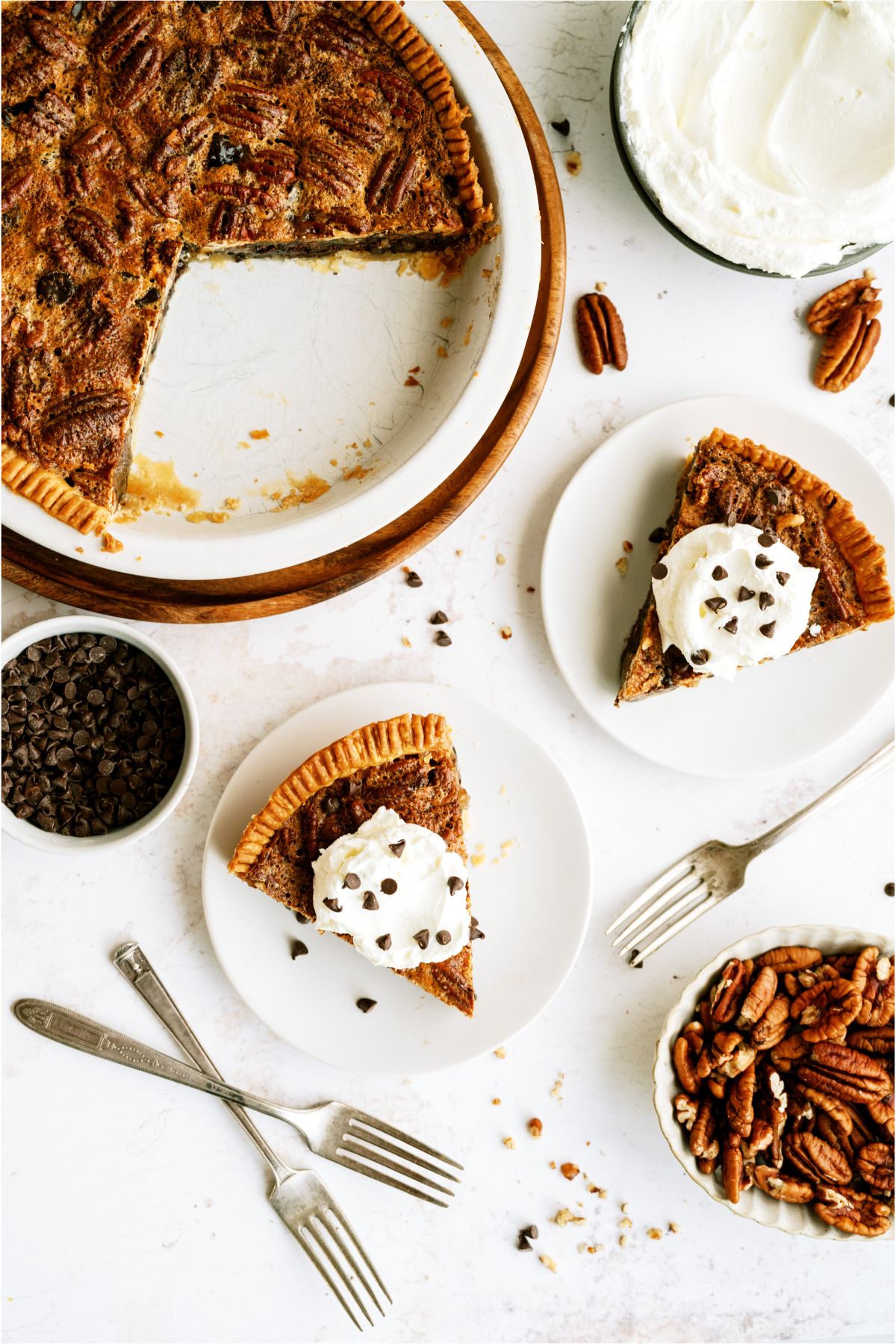 Top view of a Chocolate Pecan Pie in a pie dish with two slices in front of it, on plates topped with whip cream. Forks and a small bowl of pecans in the picture.