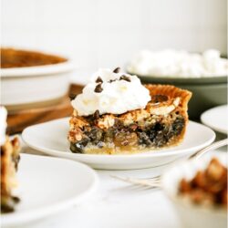 A slice of Chocolate Pecan Pie topped with whip cream on a white plate.