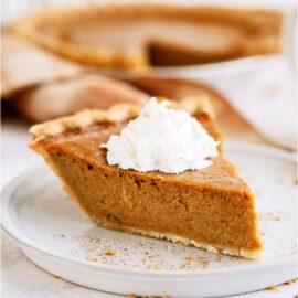 A slice of pumpkin pie topped with whip cream on a plate. With the remaining pie in the background.