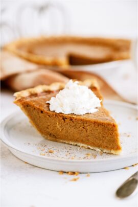 A slice of pumpkin pie topped with whip cream on a plate. With the remaining pie in the background.