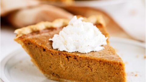 A slice of pumpkin pie topped with whip cream on a plate. With the remaining pie in the background.