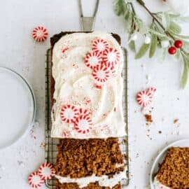 A loaf of gingerbread cake with white icing and peppermint candies, partially sliced, on a cooling rack. Nearby are scattered candies, crumbs, and a white cloth.