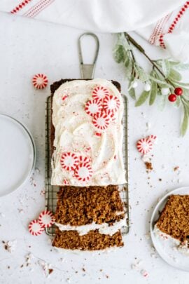 A loaf of gingerbread cake with white icing and peppermint candies, partially sliced, on a cooling rack. Nearby are scattered candies, crumbs, and a white cloth.