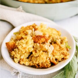 A white bowl full of Homemade Cornbread Dressing with dried herbs on the side and the remaining Cornbread dressing in the background.