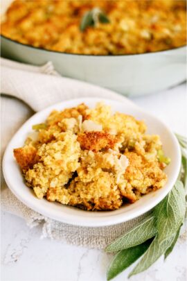 A white bowl full of Homemade Cornbread Dressing with dried herbs on the side and the remaining Cornbread dressing in the background.