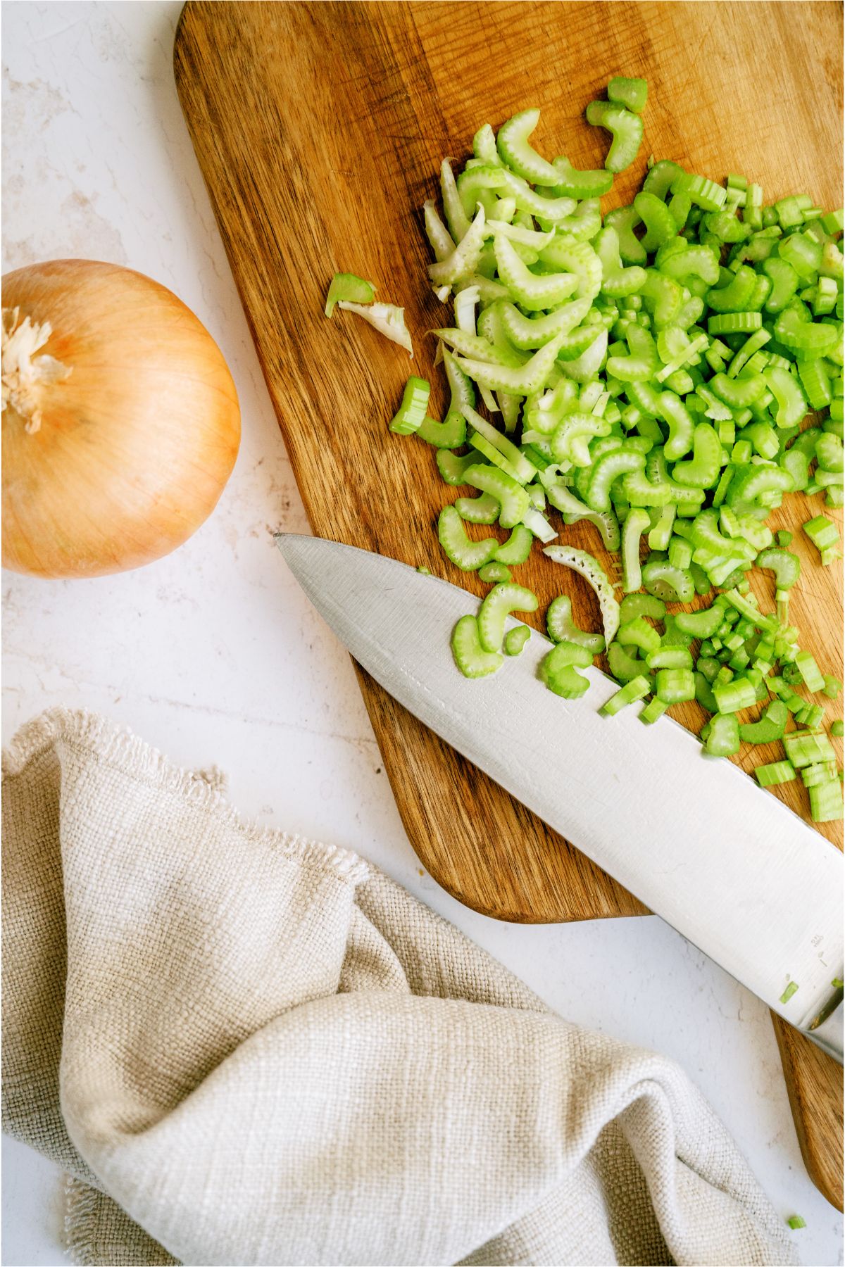 A knife next to a cutting board with diced celery and a whole onion on the side.