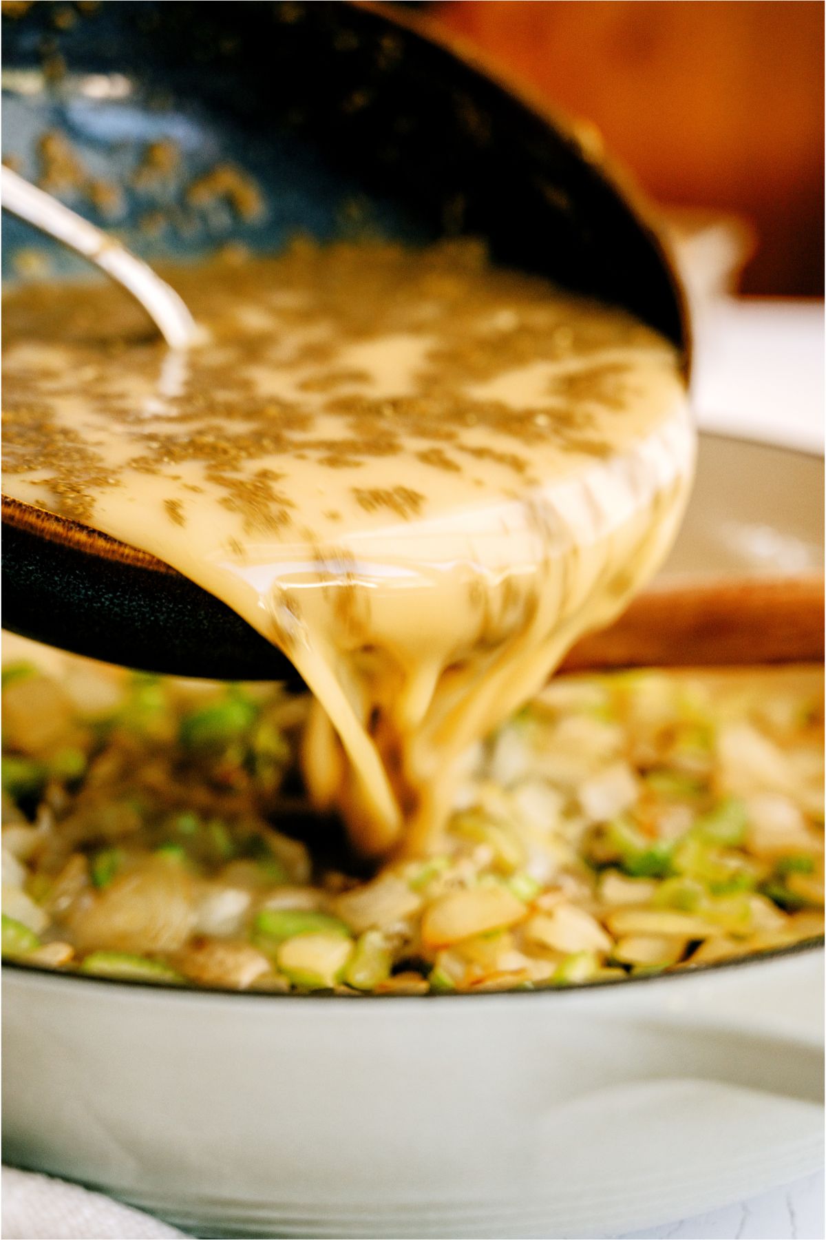 Pouring remaining sauce ingredients for cornbread dressing into skillet with celery and onions.