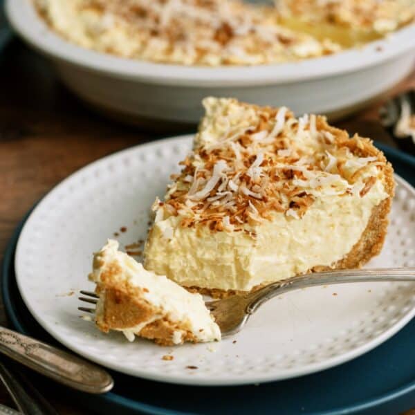 A plate with a slice of creamy, yellow pie topped with toasted coconut flakes. A fork with a bite of pie rests on the plate. The remaining pie is in the background.