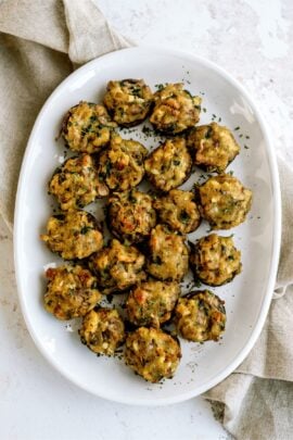 A white plate with several stuffed mushrooms topped with herbs, placed on a light surface beside a beige cloth napkin.