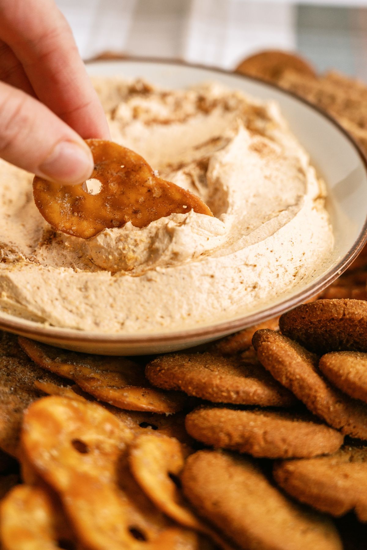 A hand dipping a cracker in a bowl of pumpkin dup with other cookies and crackers on the side.