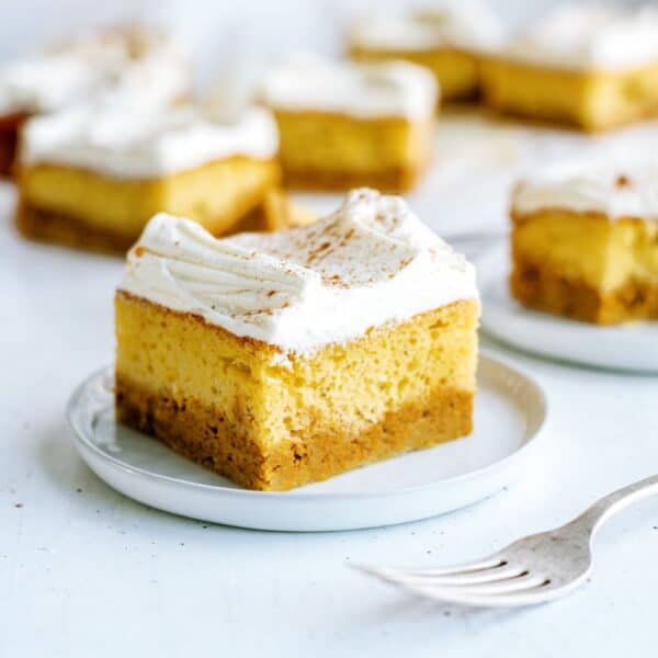 A small square pumpkin cheesecake bar with a graham cracker crust and whipped topping sits on a white plate. A fork is nearby, and similar bars are in the background.