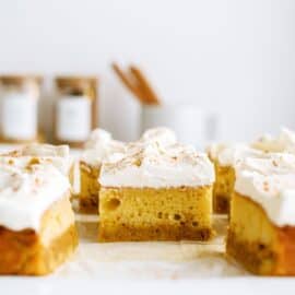 Slices of pumpkin cake topped with whipped cream and a sprinkle of cinnamon are arranged on a cutting board. Blurred containers and utensils are visible in the background.