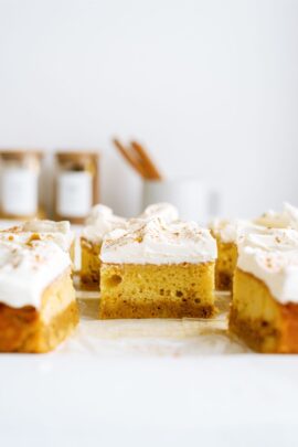 Slices of pumpkin cake topped with whipped cream and a sprinkle of cinnamon are arranged on a cutting board. Blurred containers and utensils are visible in the background.