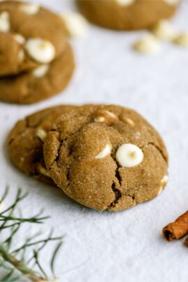 Two brown cookies with white chocolate chips on a white surface, with green sprigs partially visible.
