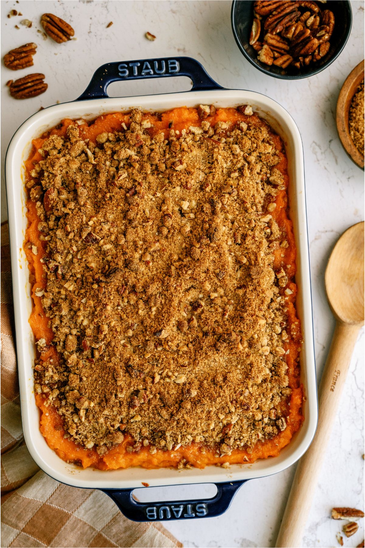 Top view of Sweet Potato Casserole in casserole dish with a wooden spoon on the side and a small dish of pecan halves in the background.