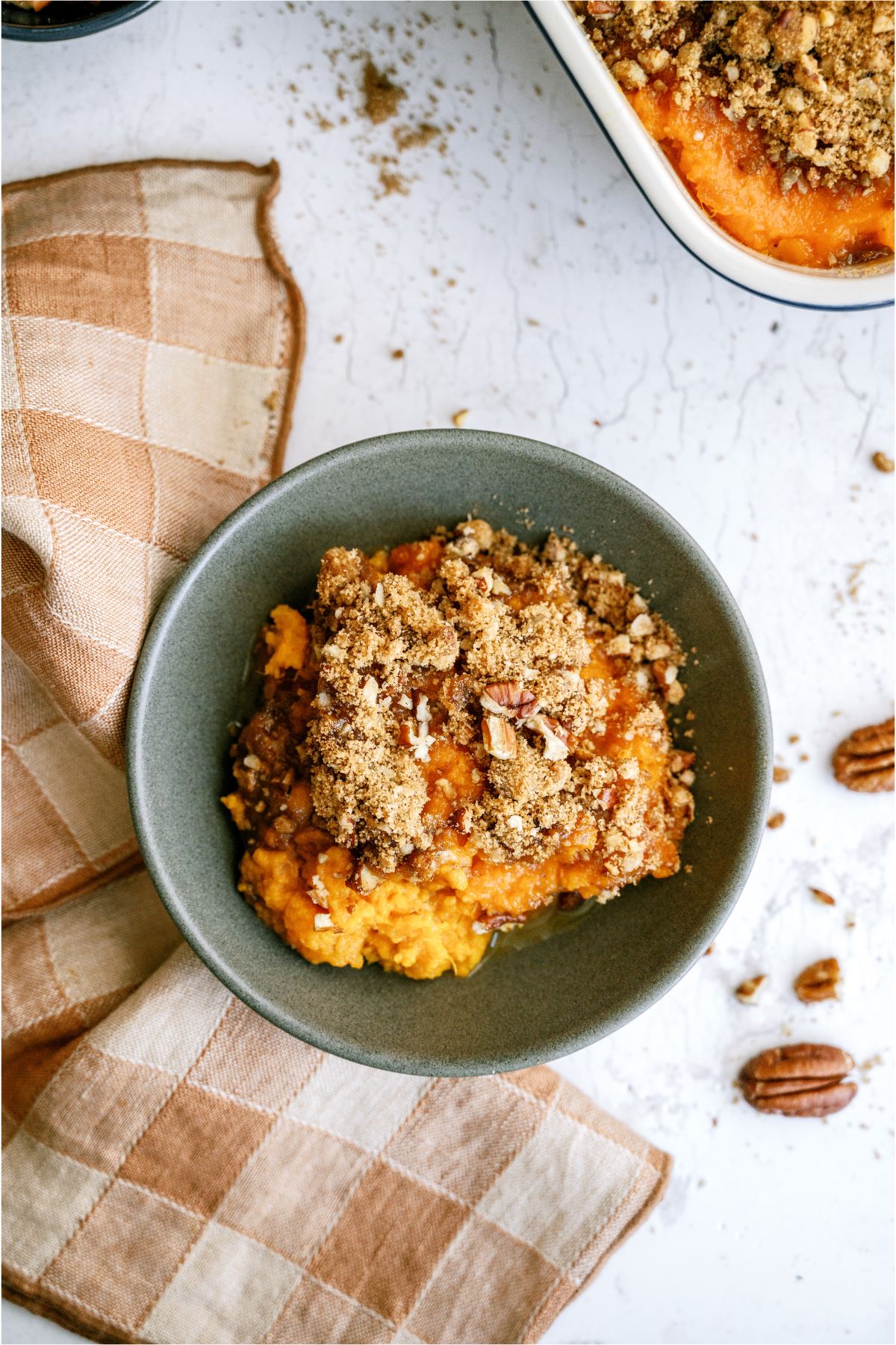 Top view of a serving of Sweet Potato Casserole in a bowl with a checkered towel underneath and the remaining Sweet Potato Casserole in the background.