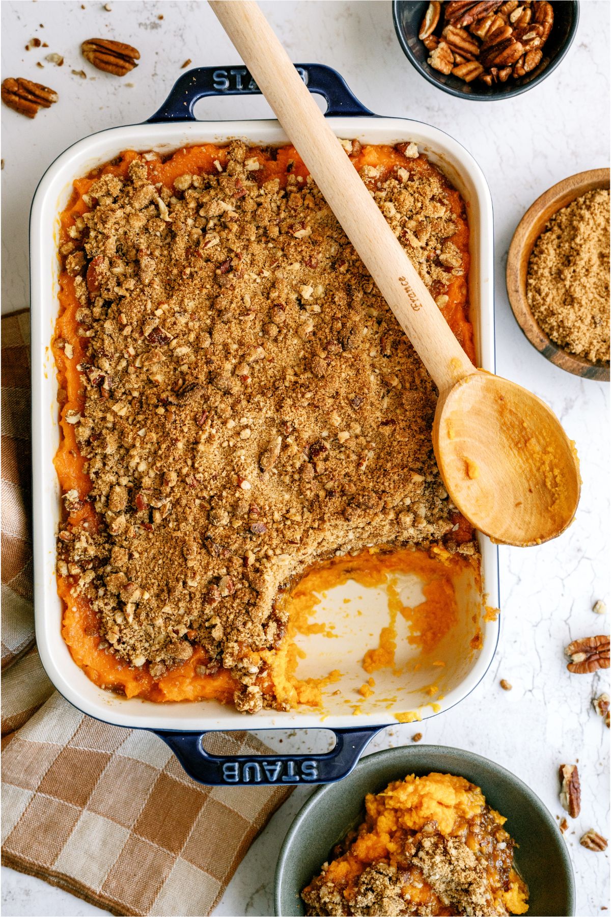 Sweet Potato Casserole  in a casserole dish with a scoop missing and a wooden spoon on top of the casserole dish. A serving of Sweet Potato Casserole in a blue bowl with two small bowl of pecans and brown sugar in the background.