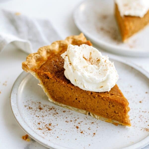 A slice of sweet potato pie topped with whipped cream on a white plate. Another slice is visible in the background.