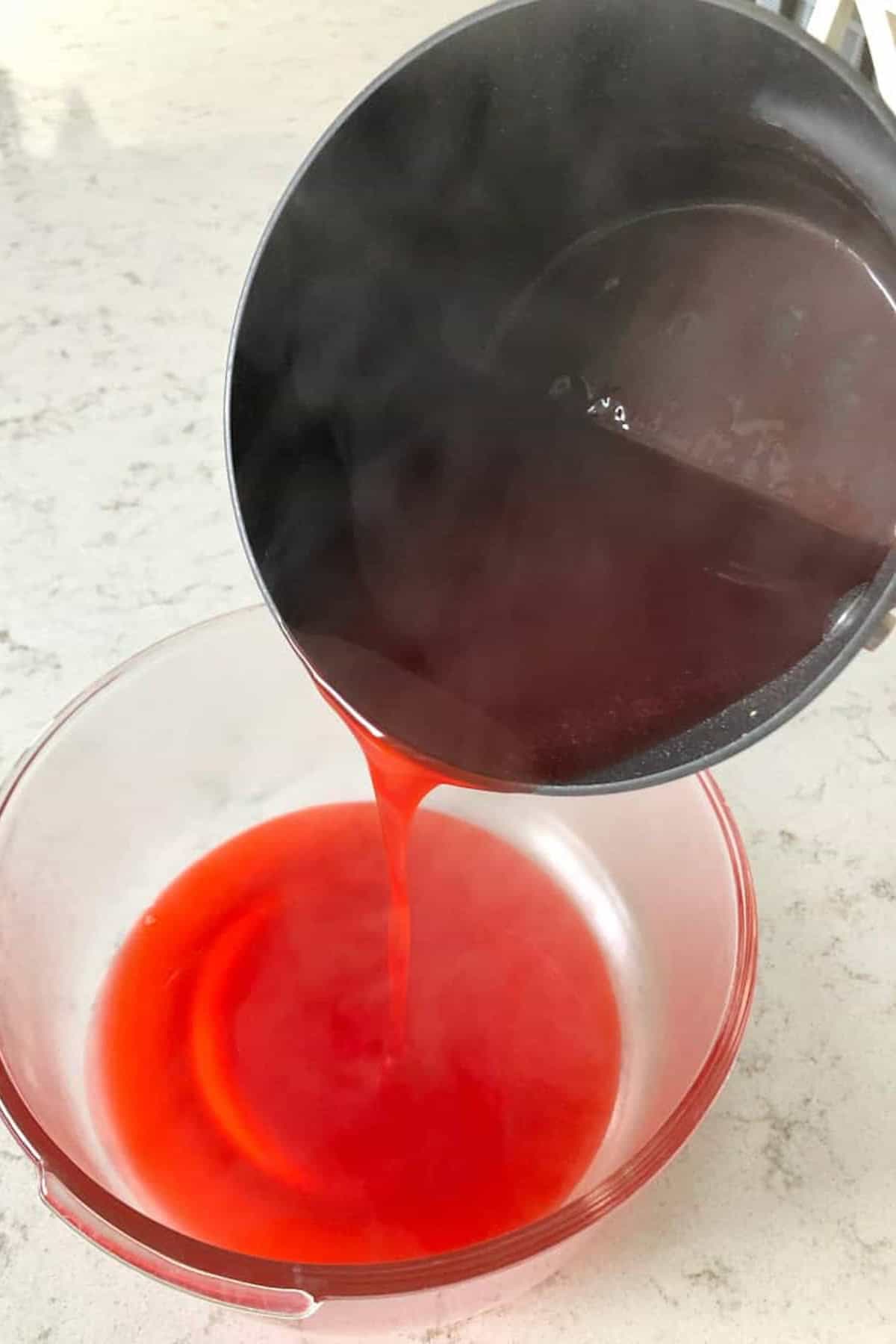 A person pours hot, red liquid from a dark pot into a transparent bowl on a light-colored countertop. Steam is visible from the liquid.