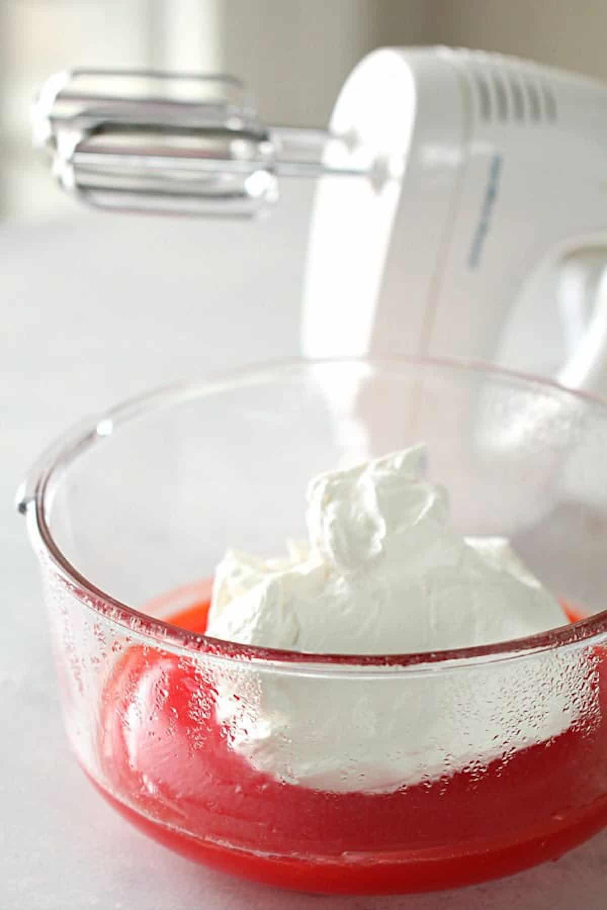 A mixing bowl containing red gelatin and a dollop of whipped cream, with a hand mixer in the background.